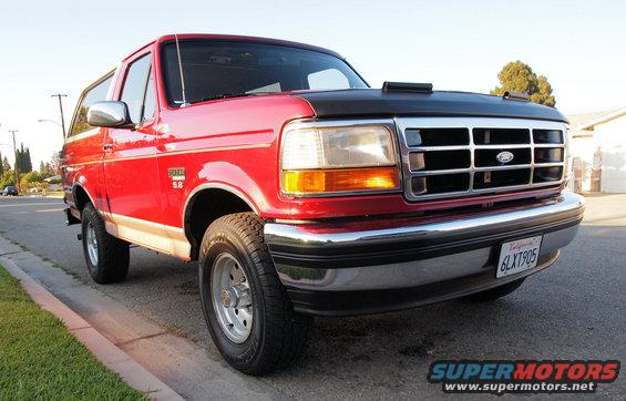 Red Ford Bronco parked on street, front quarter view. Has a black hood cover