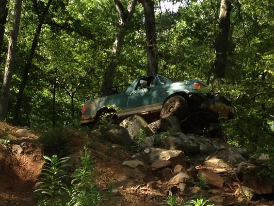 Green pickup truck on rocks, front right tire elevated. Body shows rust. Off-road tires