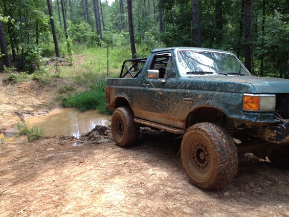 Muddy green off-road truck with roll bar, tires caked in mud, parked near a muddy puddle