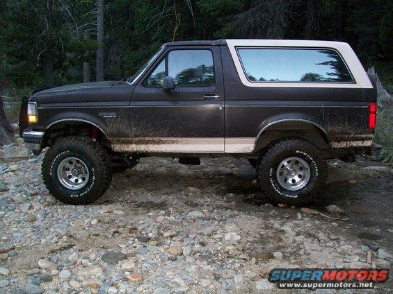 Dark brown Ford Bronco with a white top parked on a rocky surface, trees in background