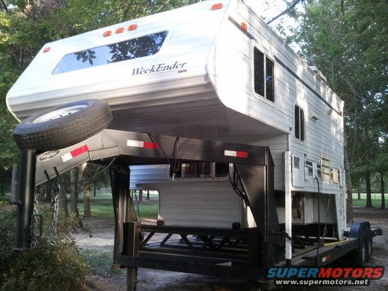 Truck camper labeled "WeekEnder" mounted on a gooseneck trailer with a spare tire. Trees in background