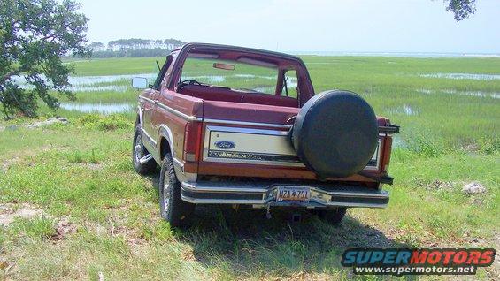 Red Ford Bronco with a spare tire on the back, parked on a grassy field near a marsh