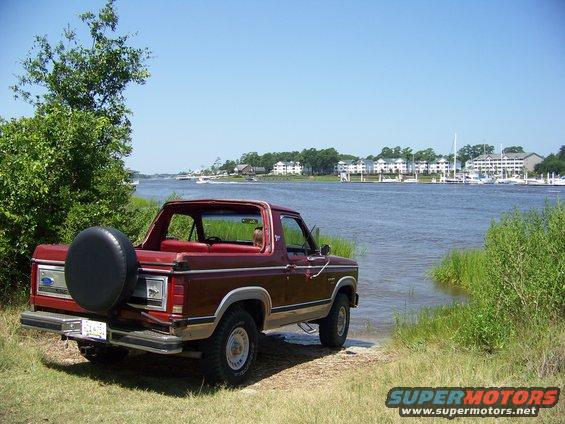 Red Ford Bronco with spare tire on back parked on grassy bank next to a wide river with buildings and boats in the distance