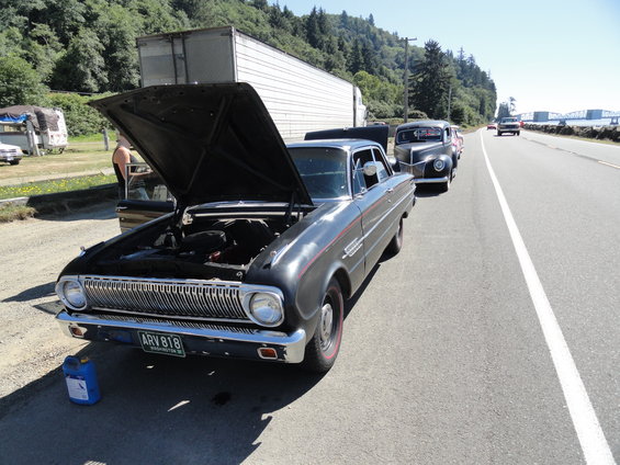 Black 1964 Ford Falcon with hood open, parked on roadside next to a trailer and other cars