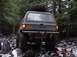 1989 Ford Bronco - An olive green SUV navigates a challenging rocky trail in a wooded area, with a spare tire mounted on its roof.