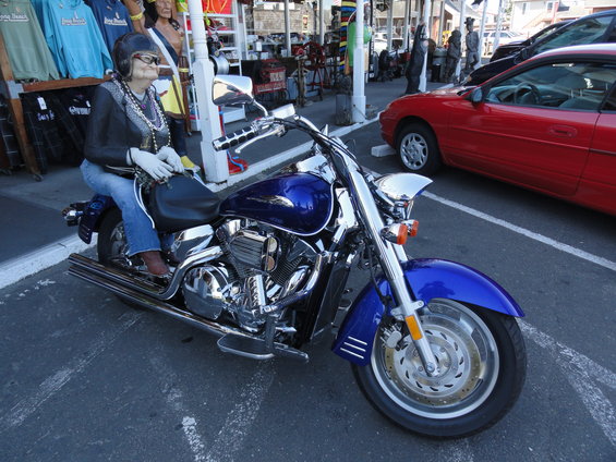 Mannequin in jeans and jacket sits on a blue motorcycle parked next to a red car