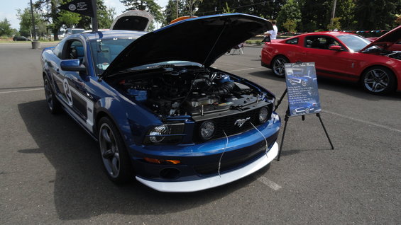 Blue Ford Mustang with hood open, red Ford Mustang in background. Display board next to blue car
