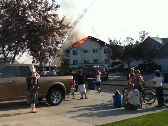 House fire with flames visible on the roof, a fire truck ladder extended, and people watching from the street