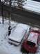 1995 Ford Probe - A white car is covered in snow on a street, with a red car parked next to it. A person is standing by the white car.