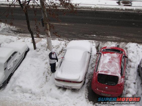 1995 Ford Probe covered in snow with a person clearing it, parked next to a red car