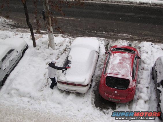 person clearing snow off white car parked next to red car in snowy lot