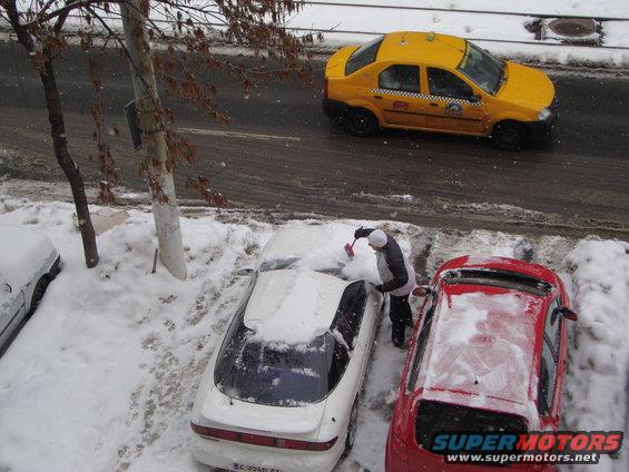 person clearing snow off white car parked next to red car, yellow taxi drives by on snowy road