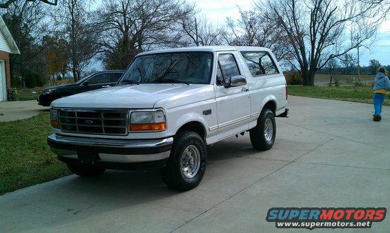 White 1995 Ford Bronco parked on a concrete driveway. A person is visible in the background on a scooter
