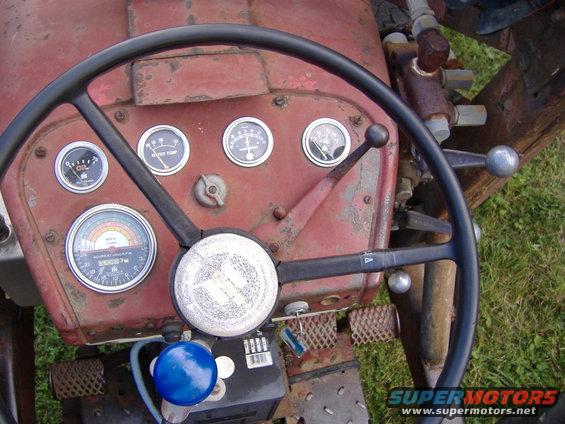 Tractor dashboard showing oil, water temp, and amp gauges, plus an hour meter. Steering wheel and control levers are visible
