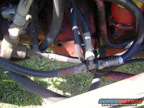 Hydraulic hoses and metal fittings on a red machine with grass in the background. "SUPERMOTORS" logo visible