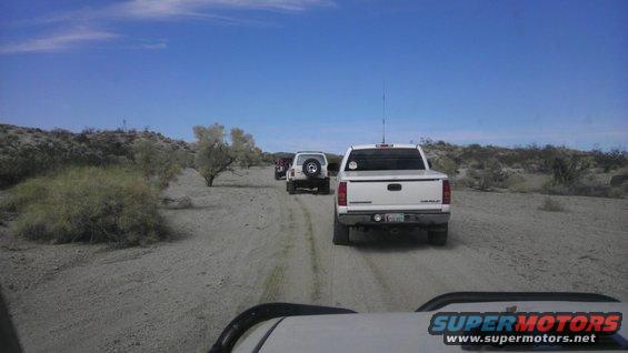 White pickup truck and two SUVs driving on a dirt road in a desert landscape