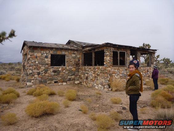 Stone building with broken windows in a desert landscape, two people standing nearby