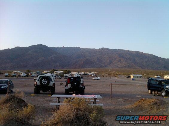 Desert landscape with parked cars, RVs, and mountains in background