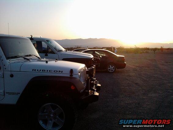 White Jeep Wrangler Rubicon in foreground, other vehicles behind, desert sunset