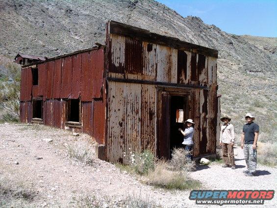 Abandoned corrugated metal building with three people standing outside in a desert landscape