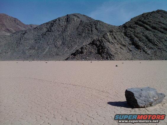 Dry lake bed with mountains in background and a rock in foreground. Supermotors logo at bottom right