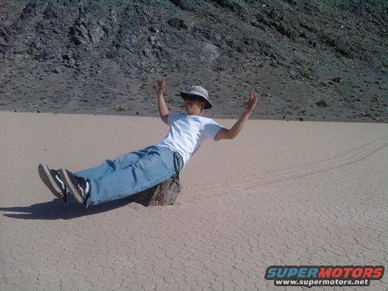 Person in hat and jeans sits on a rock in a dry, cracked desert with tire tracks visible