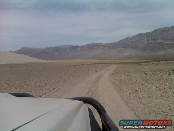 Dirt road through desert landscape with mountains in background. Part of a vehicle's roof and roll bar visible in foreground