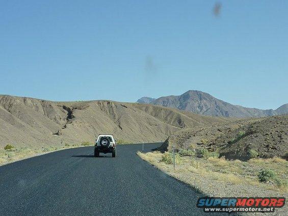 White SUV driving on a paved road through a barren, mountainous desert landscape under a clear blue sky