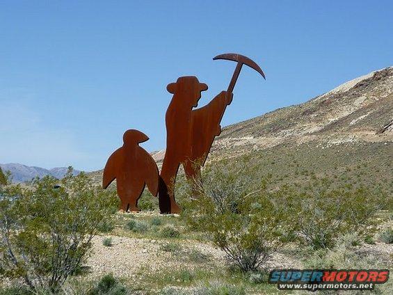 Metal sculpture of a prospector with a pickaxe and a penguin in a desert landscape
