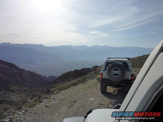 Tan SUV with spare tire on rear door drives on a dirt road with mountains in background. Another white vehicle is in the foreground