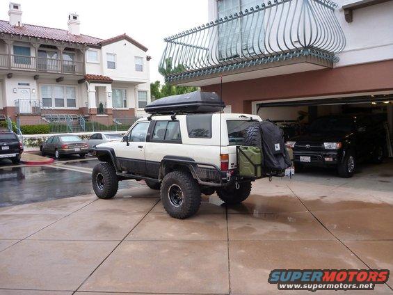 Modified white Toyota 4Runner with roof box and spare tire on back, parked in driveway next to a black SUV in a garage