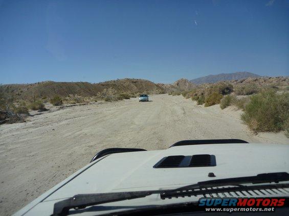 View from inside a white vehicle driving on a dirt road in a desert, with another vehicle in the distance