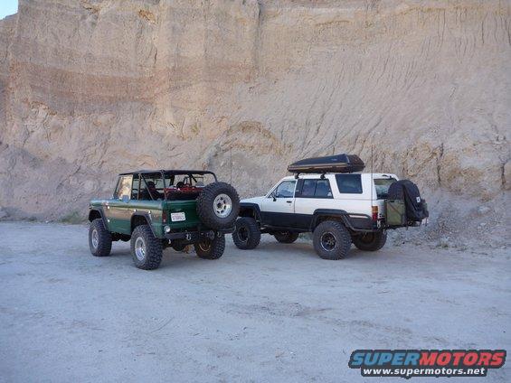 Green Ford Bronco and white Toyota 4Runner parked in desert with rocky cliff behind