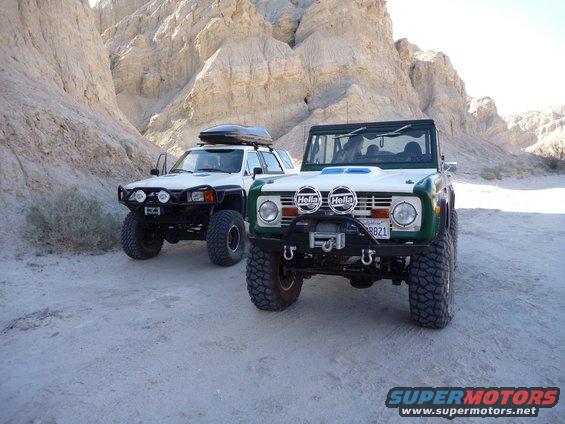 Green and white Ford Bronco with large tires and winch, parked next to a white Toyota 4Runner with a roof rack
