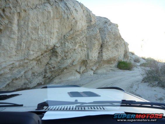 White SUV interior view driving on a sandy road beside a large rock cliff