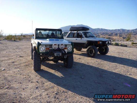 Two off-road vehicles, a classic Ford Bronco and a Toyota 4Runner with a roof box, parked on a dirt road in a desert landscape