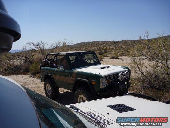 Green and white Ford Bronco with roll cage parked in desert, another vehicle in foreground