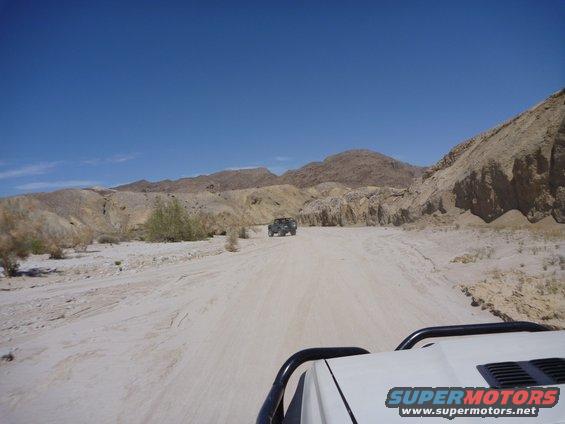 White SUV hood and roll bar in foreground, dark SUV in distance on dirt road, desert mountains under blue sky