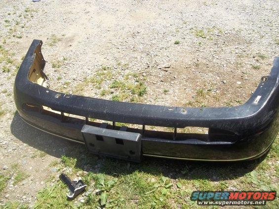 Front bumper assembly, dark colored, with license plate bracket attached. A detached bracket is on the ground