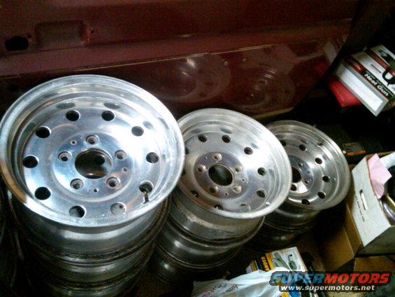 Several aluminum wheels stacked up, with a maroon vehicle body in the background