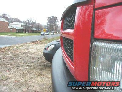 Red truck grille and headlight close-up, with a black car visible behind it on a dirt roadside