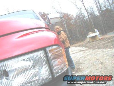 Red pickup truck front end, person leaning on it. Woods and construction in background