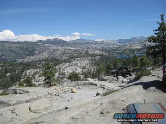 Two off-road vehicles on a rocky mountain trail with a lake in the distance