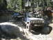 1986 Ford Bronco - Two off-road vehicles navigate a challenging, boulder-strewn path through a wooded area.