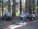 1986 Ford Bronco - Two lifted Ford Broncos are parked in a forest clearing with people standing by them.