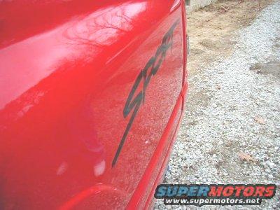 Red car side panel with black 'Sport' decal, gravel on the ground
