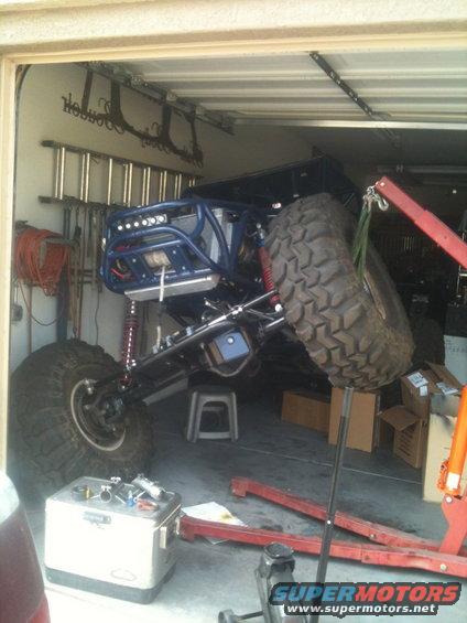 Blue off-road buggy with large tires, lifted suspension, and roll cage, partially supported by a red engine hoist in a garage