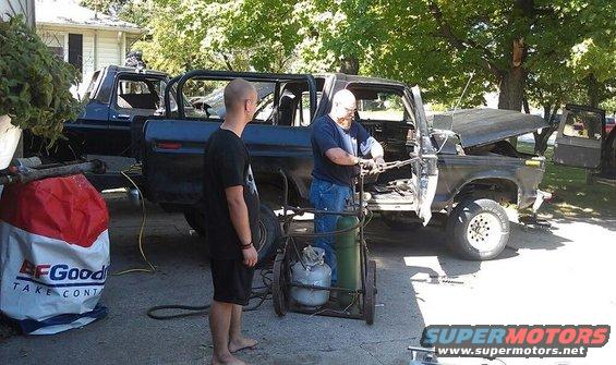 Two men are working on a dark-colored SUV that appears to be a Ford Bronco. One man is using a welding torch. Another SUV is visible in the background