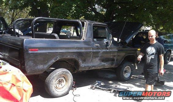 Black Ford Bronco with roll cage, hood open, and a man standing nearby. Another vehicle is partially visible behind it