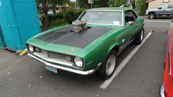 Green 1967 Camaro with a big block engine sticking out of the hood, parked next to a porta-potty